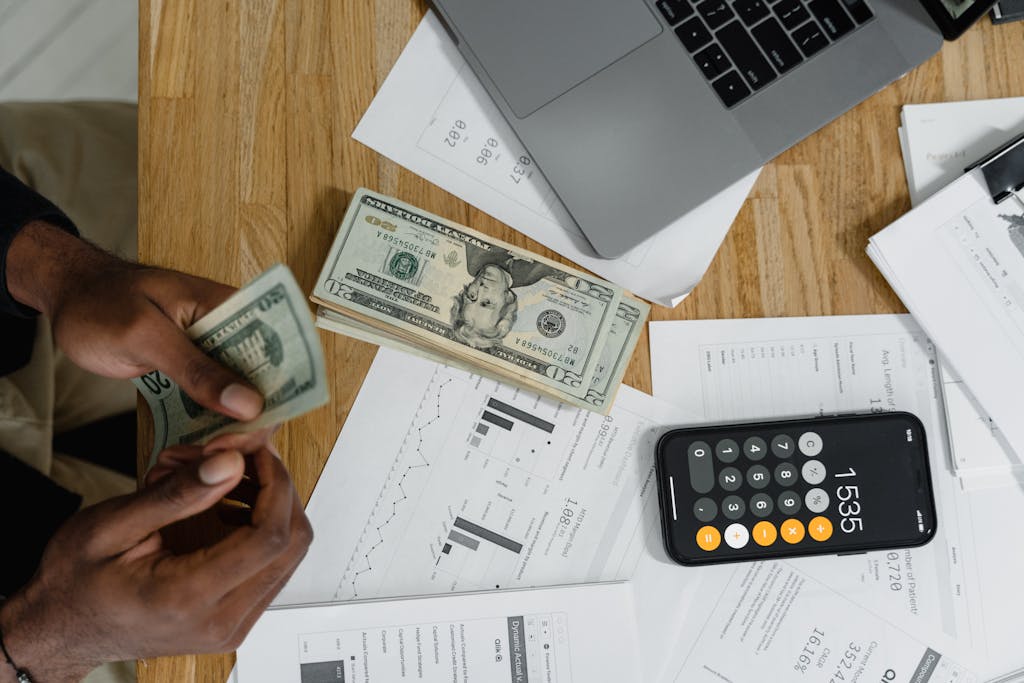 Close-up of hands counting cash on desk with calculator, charts, and laptop, illustrating financial management.